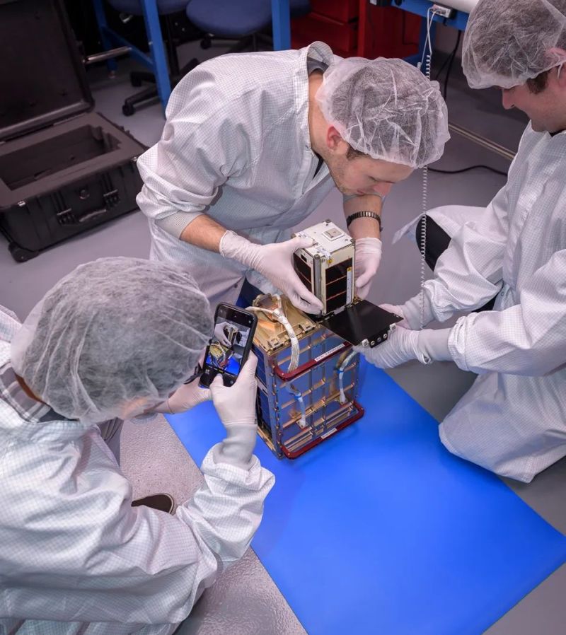 On Nov. 8, 2023, Max Holliday, middle, installs one of the four PY4 spacecraft into the dispenser supplied by Maverick Space Systems ahead of vibration testing. David Pignatelli, Maverick Space Systems, right, holds the dispenser steady as Watson Attai, left, documents the installation with a smart phone camera.